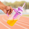 Hand holding a pink and clear bottle with a straw, filled with orange liquid, on a blurred outdoor background.