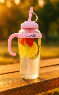 Glass water bottle with pink lid and handle, filled with fruit slices on a wooden surface.